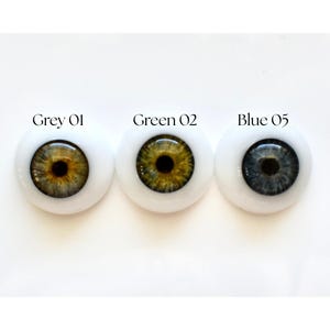 K&ouml;nnte beinhalten: Drei Puppenaugen mit unterschiedlich farbigen Iris, eingesetzt in wei&szlig;em Kunststoff. Die Iris sind grau, gr&uuml;n und blau. &Uuml;ber jedem Auge steht der Text "Grey 01", "Green 02" und "Blue 05".