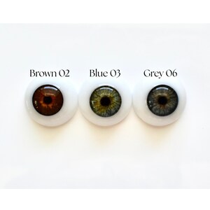 K&ouml;nnte beinhalten: Drei Puppenaugen in Braun, Blau und Grau, jeweils mit wei&szlig;em Hintergrund. Das braune Auge ist mit "Brown 02" beschriftet, das blaue Auge mit "Blue 03" und das graue Auge mit "Grey 06".