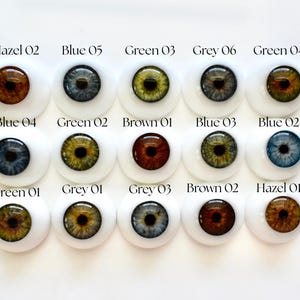 K&ouml;nnte beinhalten: Eine Sammlung von f&uuml;nfzehn Puppenaugen in verschiedenen Farben, darunter Hazel, Blau, Gr&uuml;n und Grau. Jedes Auge ist in einer wei&szlig;en, abgerundeten Basis eingesetzt, mit der Farbe und Nummer jedes Auges dar&uuml;ber gedruckt.