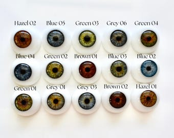 May include: A collection of fifteen doll eyes in various colors, including hazel, blue, green, and grey. Each eye is set in a white, rounded base, with the color and number of each eye printed above it.