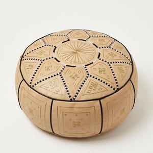 May include: A round, beige leather pouf featuring intricate gold-colored embroidery in geometric and starburst patterns. The surface is divided into panels by black piping, with alternating square and diamond shapes decorated with stitched details.