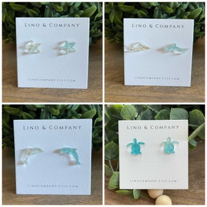 May include: Four cards with the text "LINO & COMPANY" and "LINOCOMPANY.ETSY.COM" printed on them. Each card features a pair of earrings with a different ocean-themed design: starfish, dolphins, and sea turtles. The earrings are made of clear resin with a light blue and green color gradient.