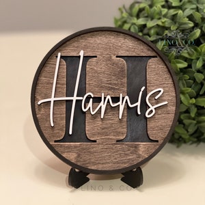 May include: A round wooden sign with a black background and a white outline. The sign says "Harris" with a large "H" in the center. The sign is on a small black stand.