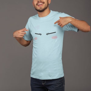 May include: A light blue t-shirt with a cartoon face printed on the front. The face has black eyebrows, pink cheeks, and a neutral expression.