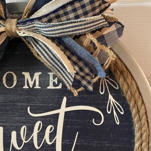 May include: Round wooden sign with a navy blue background and the words "HOME sweet" in white script. The sign is trimmed with rope and topped with a bow made of blue and tan patterned ribbons. Decorative home decor.