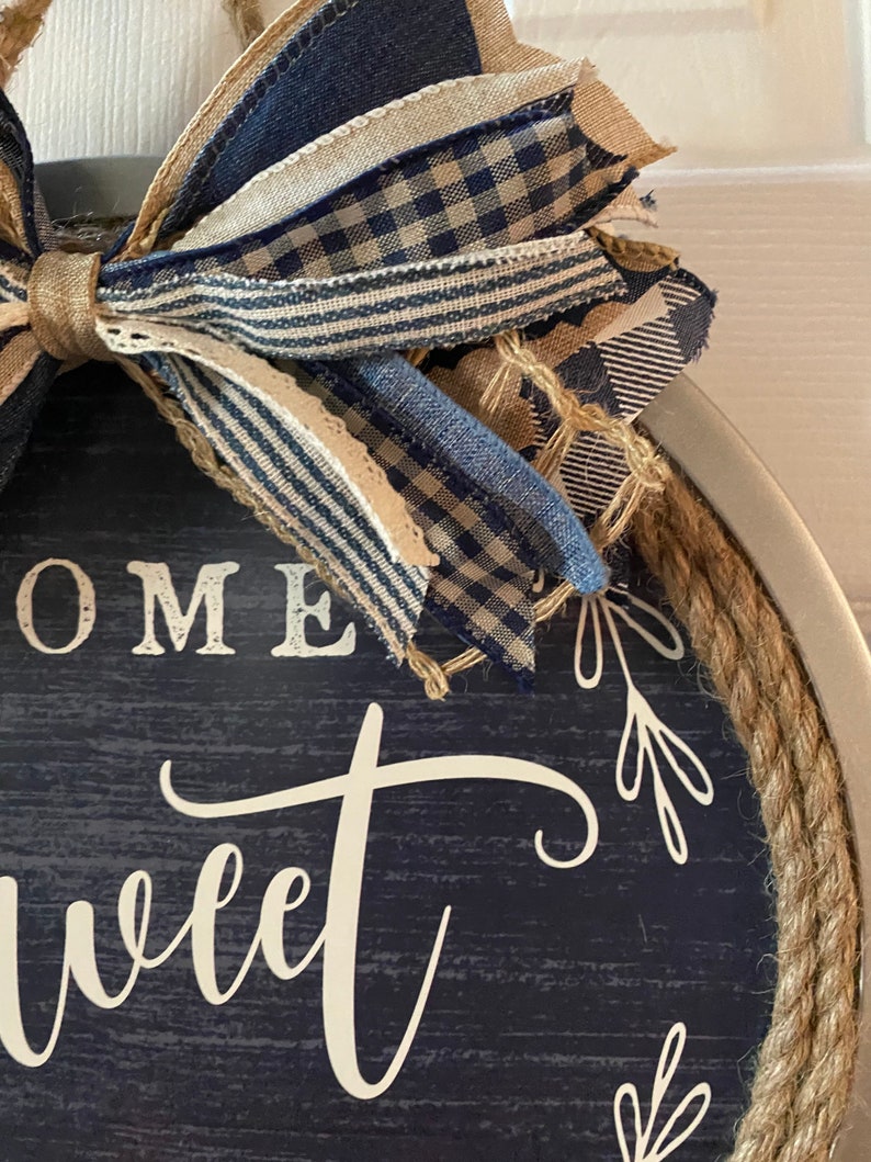 May include: Round wooden sign with a navy blue background and the words "HOME sweet" in white script. The sign is trimmed with rope and topped with a bow made of blue and tan patterned ribbons. Decorative home decor.