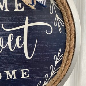 May include: Round navy blue wooden sign with the words "Home Sweet Home" in white script. The sign is trimmed with a thick rope and a silver metal frame. A bow made of burlap and blue and white plaid fabric is attached to the top.
