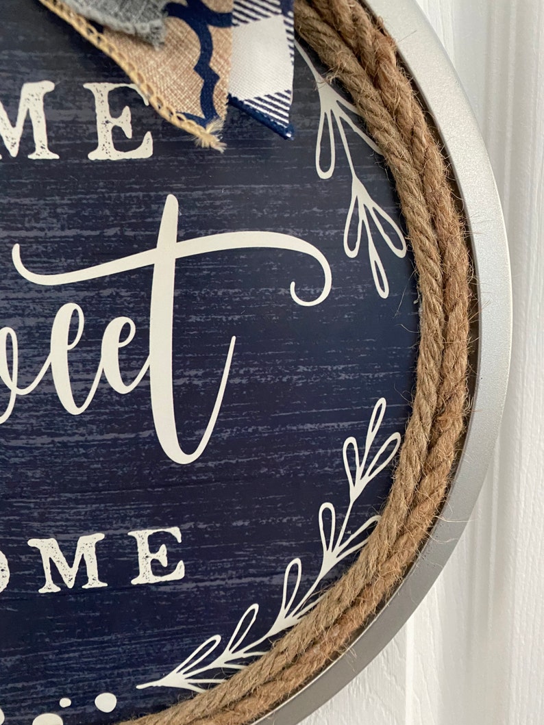 May include: Round navy blue wooden sign with the words "Home Sweet Home" in white script. The sign is trimmed with a thick rope and a silver metal frame. A bow made of burlap and blue and white plaid fabric is attached to the top.