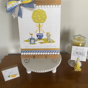 May include: A white wooden sign with a lemon tree illustration and the text "When life gives you lemons, make lemonade". The sign is on a white stand with a gold metal base. There is a small sign that says "Easy peasy lemon squeezy" and a sign that says "Ice Cold Lemonade 5¢".