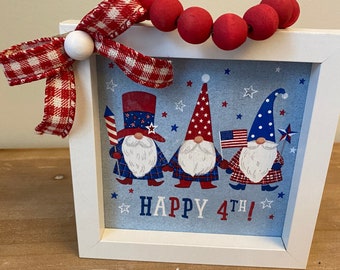 May include: A white framed picture with three gnomes wearing red, white, and blue hats and outfits. The gnomes are holding flags and the picture says "HAPPY 4TH!" A red and white checkered bow with a white bead is on top of the frame.