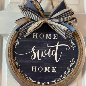 May include: Round wall hanging with a navy blue background and the words "HOME SWEET HOME" in white script. The sign is trimmed with rope and a decorative bow in shades of blue and tan.