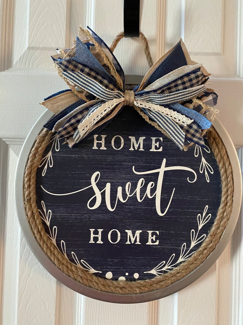 May include: Round wall hanging with a navy blue background and the words "HOME SWEET HOME" in white script. The sign is trimmed with rope and a decorative bow in shades of blue and tan.