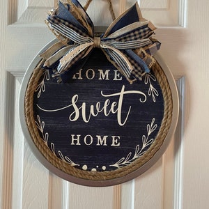 May include: Round, navy blue and silver metal wall decor with the words "HOME sweet HOME" in white script. The sign is trimmed with rope and topped with a bow made of blue and tan ribbons. A decorative piece for the home.