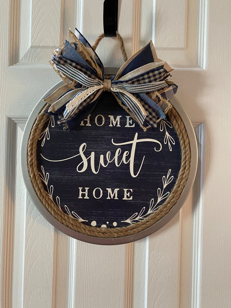 May include: Round, navy blue and silver metal wall decor with the words "HOME sweet HOME" in white script. The sign is trimmed with rope and topped with a bow made of blue and tan ribbons. A decorative piece for the home.