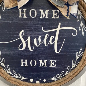 May include: Round navy blue wooden sign with the words "HOME SWEET HOME" in white script. The sign is trimmed with rope and a decorative bow made of burlap and patterned fabric.