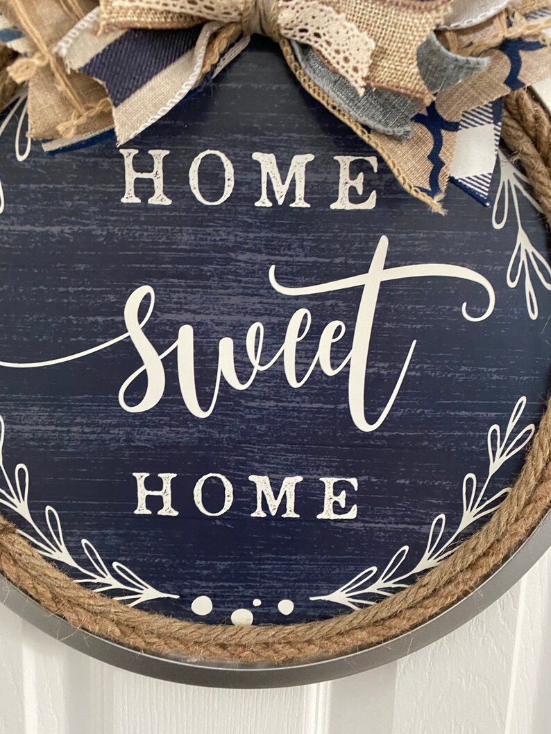 May include: Round navy blue wooden sign with the words "HOME SWEET HOME" in white script. The sign is trimmed with rope and a decorative bow made of burlap and patterned fabric.