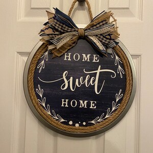 May include: Round wooden sign with a navy blue background and the words "HOME SWEET HOME" in white script. The sign is framed with a metal rim and rope, and topped with a decorative bow made of blue and tan fabric.