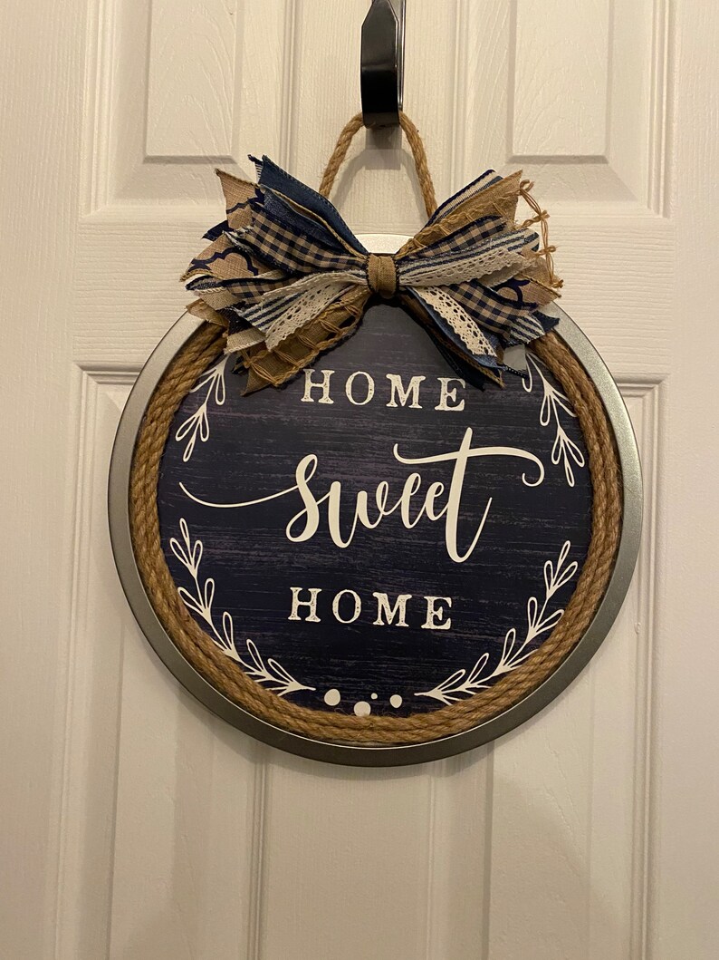 May include: Round wooden sign with a navy blue background and the words "HOME SWEET HOME" in white script. The sign is framed with a metal rim and rope, and topped with a decorative bow made of blue and tan fabric.