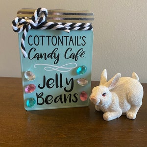 May include: A light blue sign with the text "Cottontail's Candy Cafe" and "Jelly Beans" in black lettering. The sign is decorated with colorful jelly beans. A white rabbit figurine is sitting next to the sign.