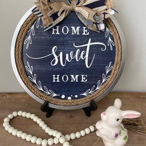 May include: A round, navy blue wooden sign with the words "HOME SWEET HOME" in white script. The sign has a rope border and a decorative bow at the top. A white ceramic bunny and a beaded garland are in front of the sign.