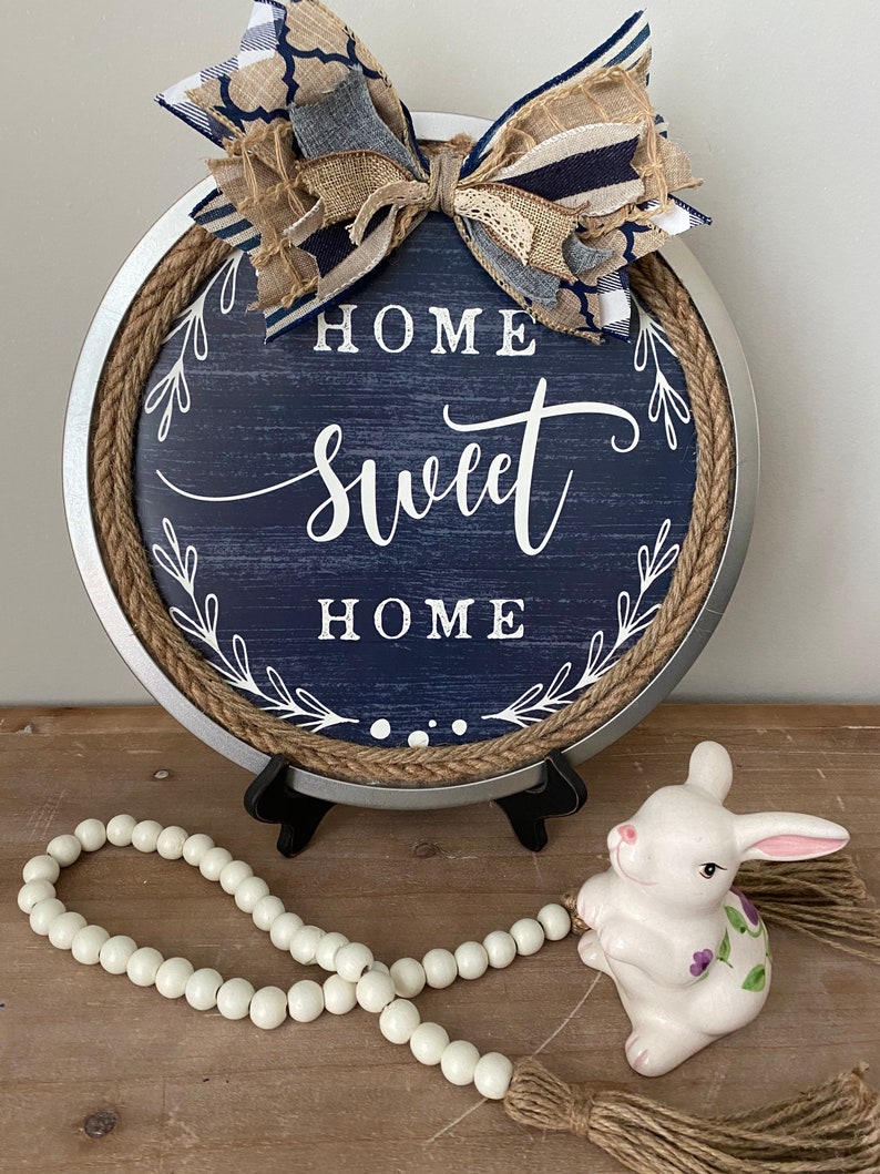 May include: A round, navy blue wooden sign with the words "HOME SWEET HOME" in white script. The sign has a rope border and a decorative bow at the top. A white ceramic bunny and a beaded garland are in front of the sign.