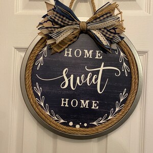 May include: Round navy blue and silver metal sign with the words "HOME SWEET HOME" in white script. The sign has a rope border and a decorative bow made of blue and tan fabric.