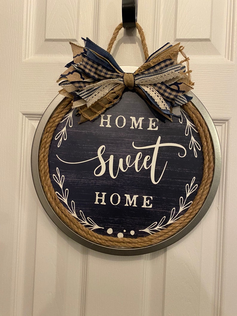 May include: Round navy blue and silver metal sign with the words "HOME SWEET HOME" in white script. The sign has a rope border and a decorative bow made of blue and tan fabric.