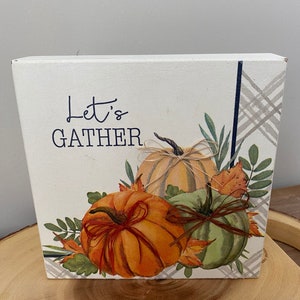 May include: A white wooden sign with the text "Let's Gather" and a painting of three pumpkins with fall leaves. The pumpkins are orange, green, and beige.