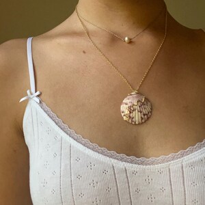May include: A two-strand gold necklace with a pearl pendant and a seashell pendant. The seashell is pink, purple, and cream-colored. The person is wearing a white tank top with a lace trim.