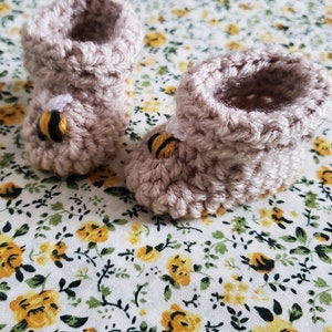Puede incluir: Un par de botines de bebé de punto beige con un diseño de abeja en cada botín. Los botines están sobre un fondo blanco con un patrón floral amarillo.