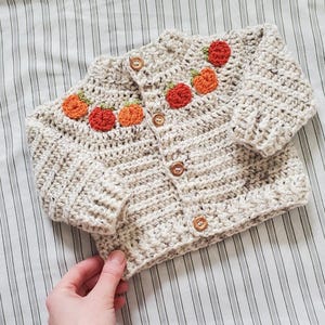 May include: A cream-colored crocheted baby cardigan with a button closure and orange embroidered pumpkins on the front.
