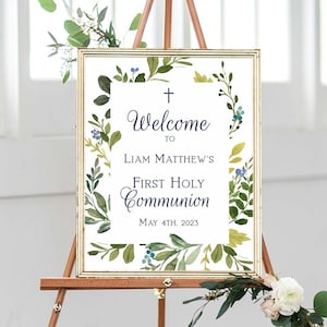 May include: Welcome to Liam Matthew's First Holy Communion sign with a gold frame and green and blue floral design. The sign is on a wooden easel with greenery and white flowers.
