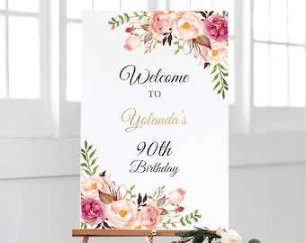 May include: A rectangular white welcome sign for a 90th birthday celebration featuring watercolor floral arrangements in shades of pink, magenta, and green. The text reads Welcome TO Yolanda's 90th Birthday in black and gold script and serif fonts.