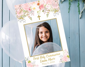 May include: A white photo booth prop with a gold frame and a floral design. The frame has a cross at the top and the text "First Holy Communion Emma Marie May 11, 2019".