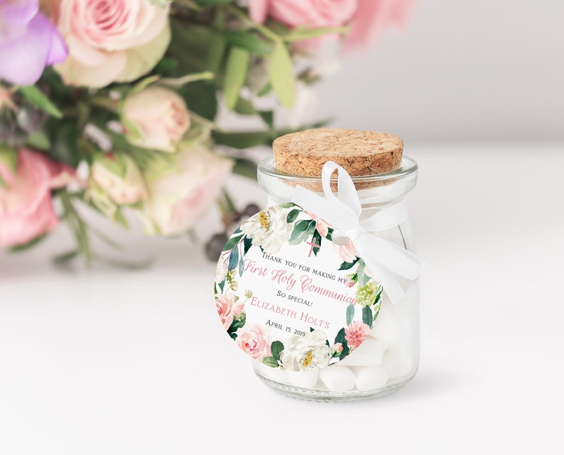 Pode incluir: Um pequeno frasco de vidro com tampa de corti&ccedil;a cheio de doces brancos. Uma etiqueta redonda branca com desenhos florais rosa e verdes est&aacute; presa ao frasco com uma fita branca. A etiqueta diz "Obrigado por tornar a minha Primeira Comunh&atilde;o t&atilde;o especial! Elizabeth Holt's Abril 15, 2019".