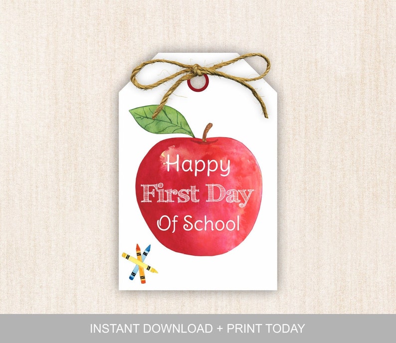 May include: A white gift tag with a red apple illustration and the text "Happy First Day of School". There are three colorful crayons in the bottom left corner of the tag.
