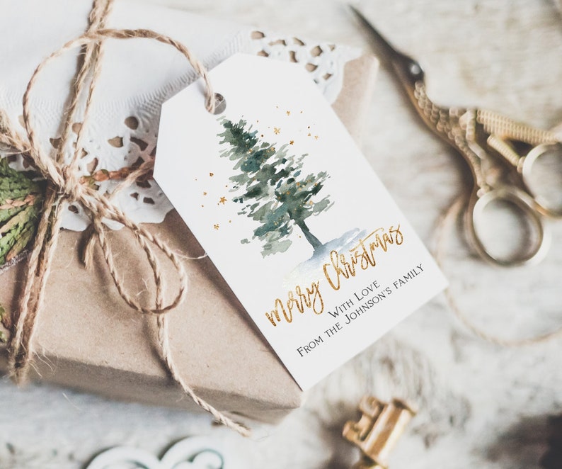 May include: A white gift tag with a watercolor illustration of a green pine tree with gold glitter. The text "merry christmas" is written in gold script with the words "with love from the Johnson's family" below.