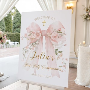May include: A white sign on an easel with the text "Welcome to Julia's First Holy Communion April 20th 2026." The sign features a watercolor illustration of a pink bow with floral accents and a gold cross.