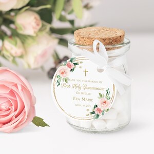 May include: A small glass jar with a cork lid filled with white candy. A white ribbon is tied around the jar with a round tag attached. The tag is white with a gold border and features a floral design with pink flowers and green leaves. The tag also includes the text "Thank you for making my First Holy Communion So Special! Eva Marie April 15, 2019".