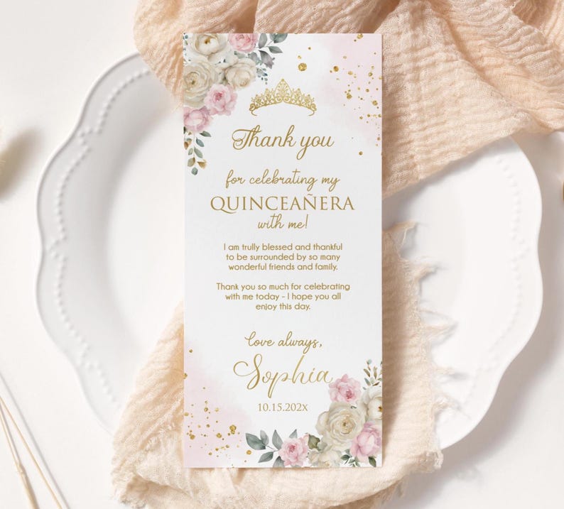 May include: A rectangular invitation card with a pink and gold design. The card features floral accents, a gold tiara, and the text "Thank you for celebrating my Quincea&ntilde;era with me!" The card is on a white plate and a beige cloth.
