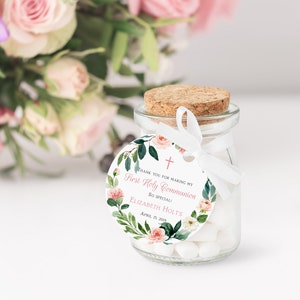 May include: A small glass jar with a cork lid and a white ribbon tied around it. The jar is filled with white candy. A round white tag with a pink floral design and the text "Thank you for making my First Holy Communion so special! Elizabeth Holt's April 13, 2019" is attached to the jar.