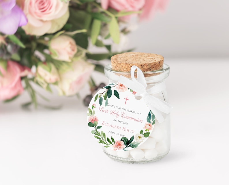 May include: A small glass jar with a cork lid and a white ribbon tied around it. The jar is filled with white candy. A round white tag with a pink floral design and the text "Thank you for making my First Holy Communion so special! Elizabeth Holt's April 13, 2019" is attached to the jar.