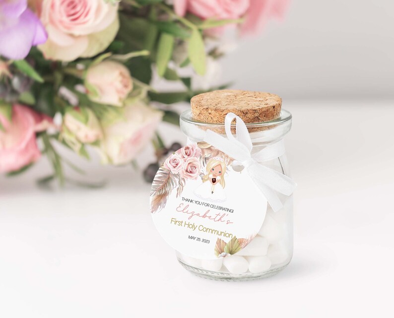 May include: A small glass jar with a cork lid and a white ribbon tied around it. The jar is filled with white candy. A round tag with a floral design and the text "Thank you for celebrating Elizabeth's First Holy Communion May 25, 2021" is attached to the ribbon.