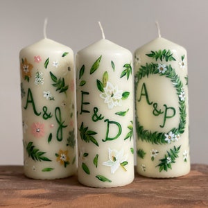 May include: Three white pillar candles with hand-painted floral designs and initials. The candles are decorated with green leaves, white flowers, and small white dots. The initials on the candles are "A & J", "E & D", and "A & P".