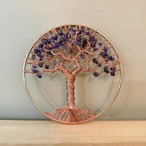 Könnte beinhalten: Handgefertigte Baumskulptur aus Kupferdraht mit kreisförmigem Rahmen. Die Äste des Baumes sind mit kleinen, violetten Amethyststeinen verziert. Der Baumstamm ist aufwendig gewebt. Das Kunstwerk ist auf einer Holzoberfläche montiert.