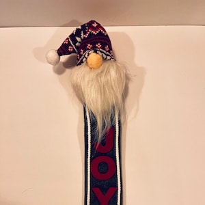 May include: A gray and white gnome with a red and white knit hat and a white beard. The gnome is holding a sign that says "JOY" in red letters.
