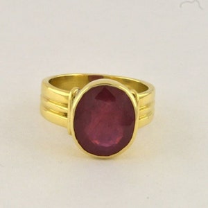 May include: A gold ring with a large oval-shaped ruby gemstone. The ring has a simple band with three grooves.