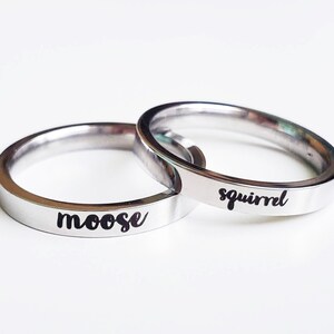 以下が含まれることがあります： 「moose」と「squirrel」の文字が黒い筆記体で刻まれた2つのシルバーカラーのリング。リングはシンプルなバンドで、ステンレス鋼などの金属製である可能性が高い。背景はプレーンな白い表面。