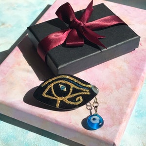 Peut inclure: Un pendentif noir avec un motif doré de l'Œil d'Horus et un charme de perle de verre bleu. Le pendentif est posé sur une surface à motifs roses et blancs, à côté d'une boîte cadeau noire nouée d'un ruban bordeaux.