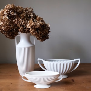 Pode incluir: Natureza morta com um vaso branco com flores secas e duas tigelas decorativas brancas com alças. O vaso tem um design canelado e as tigelas um padrão canelado. Os objetos estão sobre uma superfície de madeira.